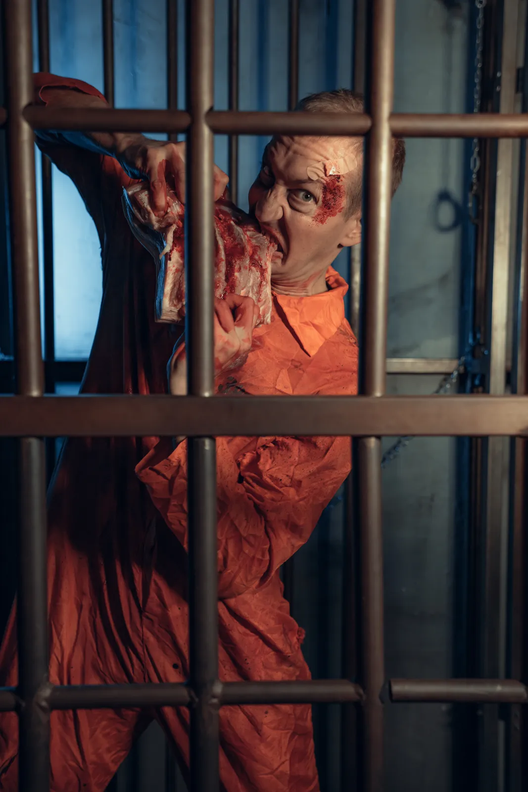 Détenu zombie agrippé aux barreaux, visage lacéré, combinaison orange, Escape Game d'Halloween