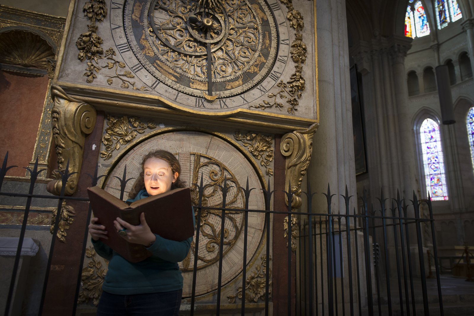 Une conteuse de Cybèle lit un grimoire à la lumière d'une lampe devant une horloge ancienne